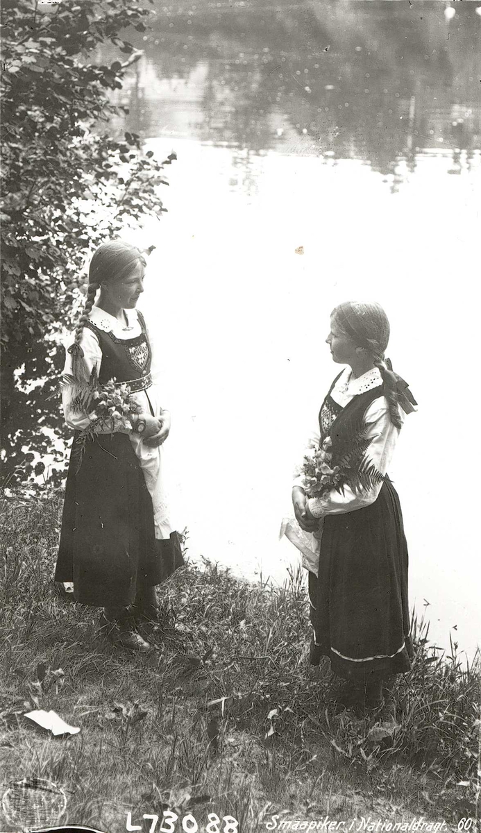 Fotografi av to jenter med blomsterbuketter, stående ved vann.  Drakt fra Hardanger