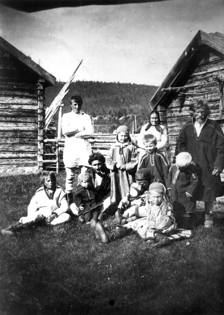Barn og voksne samer i hverdagsklær fotografert på tunet mellom to hus. Karasjok august 1897.