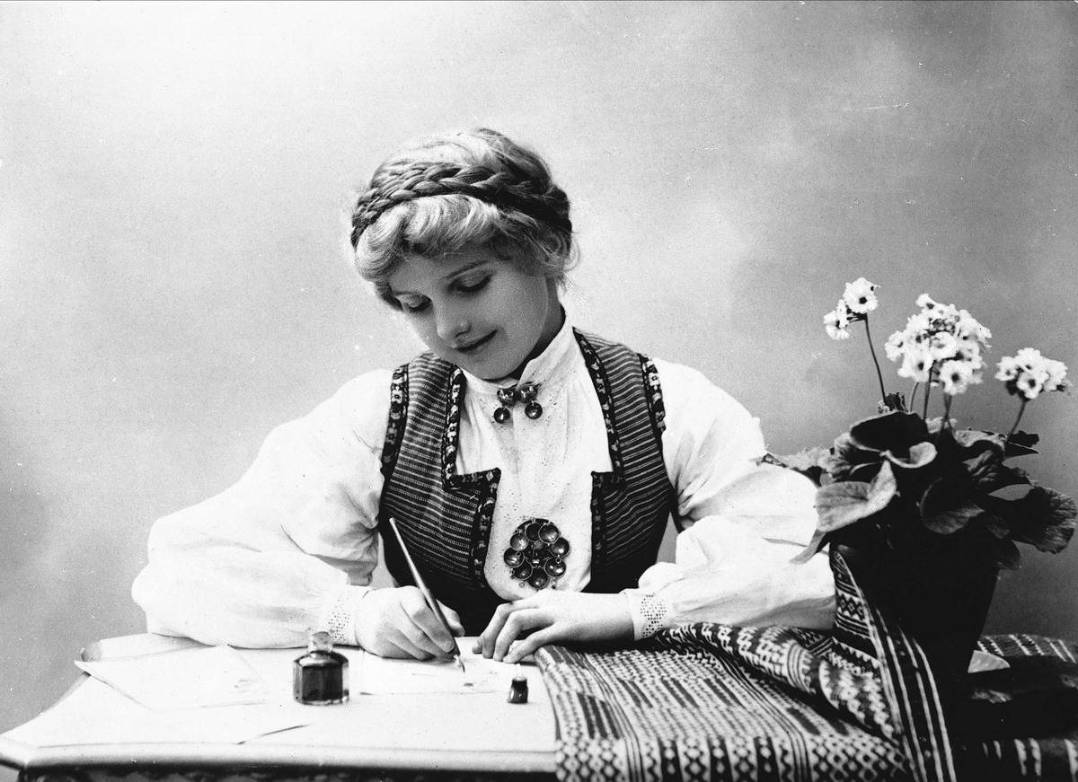 Studioportrett av kvinne i drakt med oppsatt hår. Hun sitter ved et bord og skriver et brev med penn og blekk. På bordet er det duk og blomsterpotte.