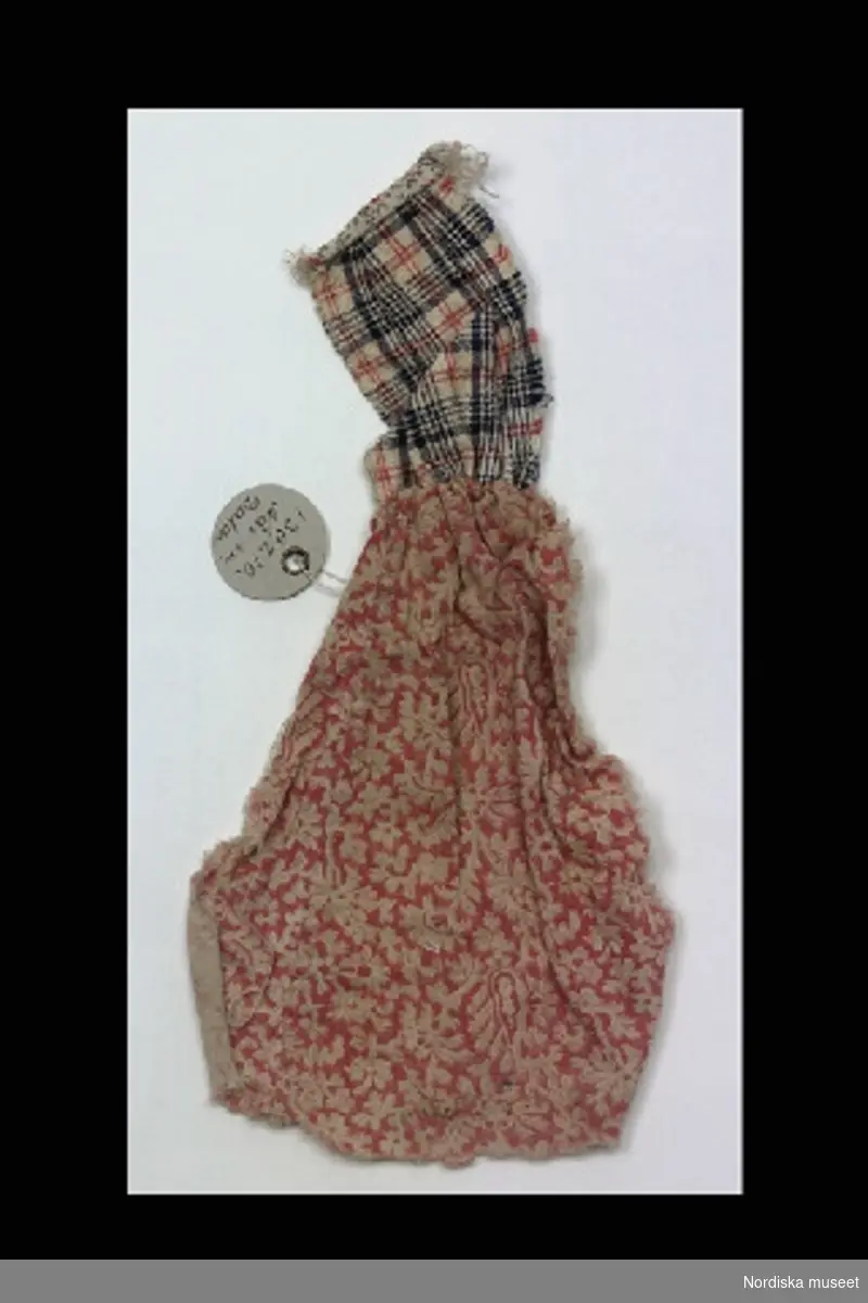 Inventering Sesam 1996-1999:
L 22,5 cm
Dockförkläde av två ihopsydda tygstycken. En större av tryckt bomull, rosa och vit. En mindre, bröstlapp, av rutigt ylletyg, vit, blå och röd. 
Leif Wallin dec 1996

