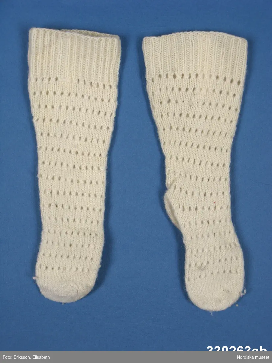 Sockor [a-b], vita långskaftade för 1-2-årig flicka. Slätstickad tå och sula, hålmönstrat skaft samt resårstickad mudd.
Anm: Något fläckig under foten.
/Zingoalla Rosenqvist 2010-04-08