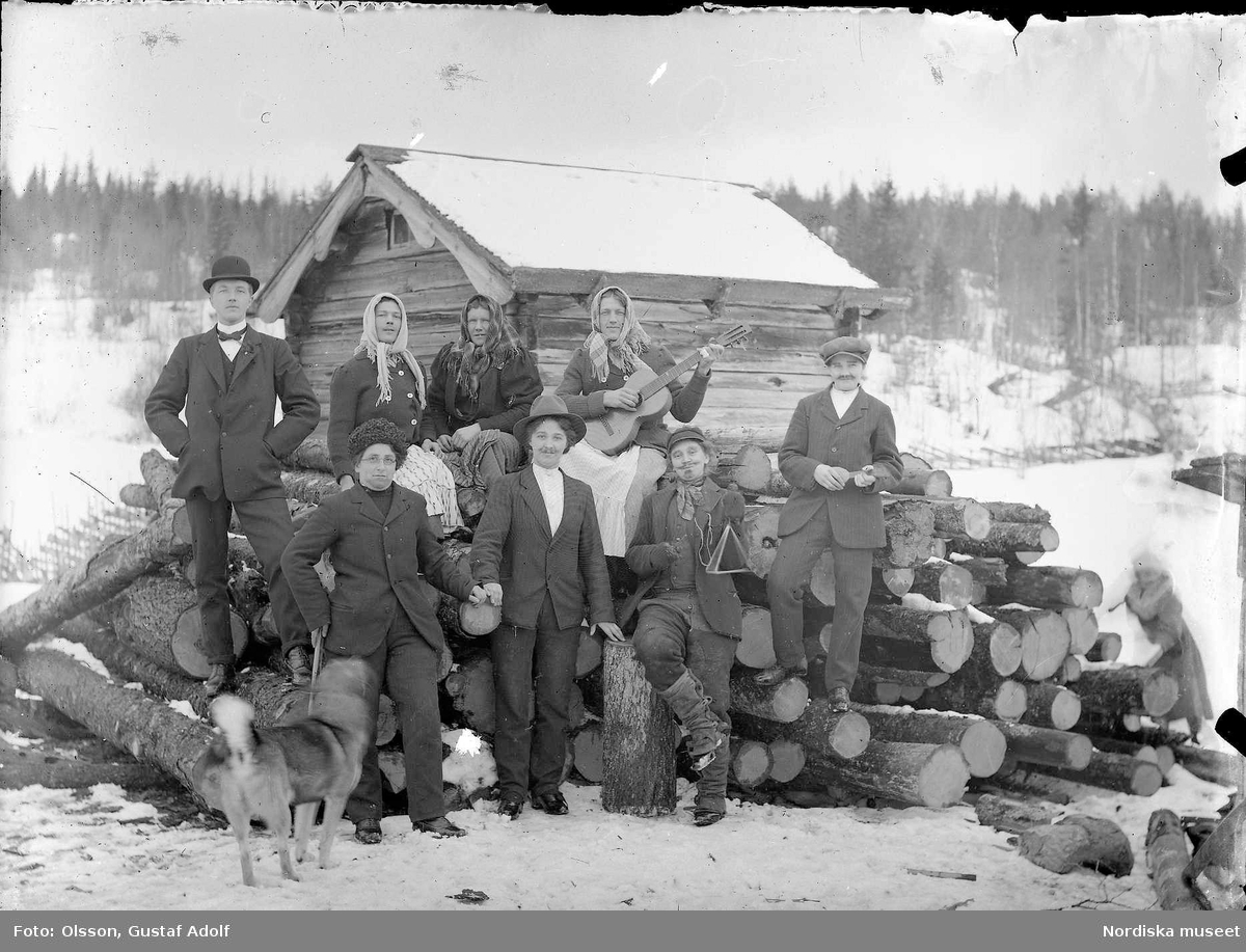 Gruppfoto med ett utklätt sällskap framför en timmerhög och en byggnad på vintern. Kvinnor klädda som män och män klädda som kvinnor. Från början av 1900-talet.