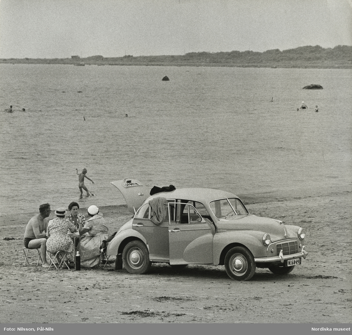 En bil, Morris Minor, parkerad på badstrand vid Frösakull, Halland. Tre kvinnor och en man sitter vid picknickbord, badande pojke i bakgrunden.