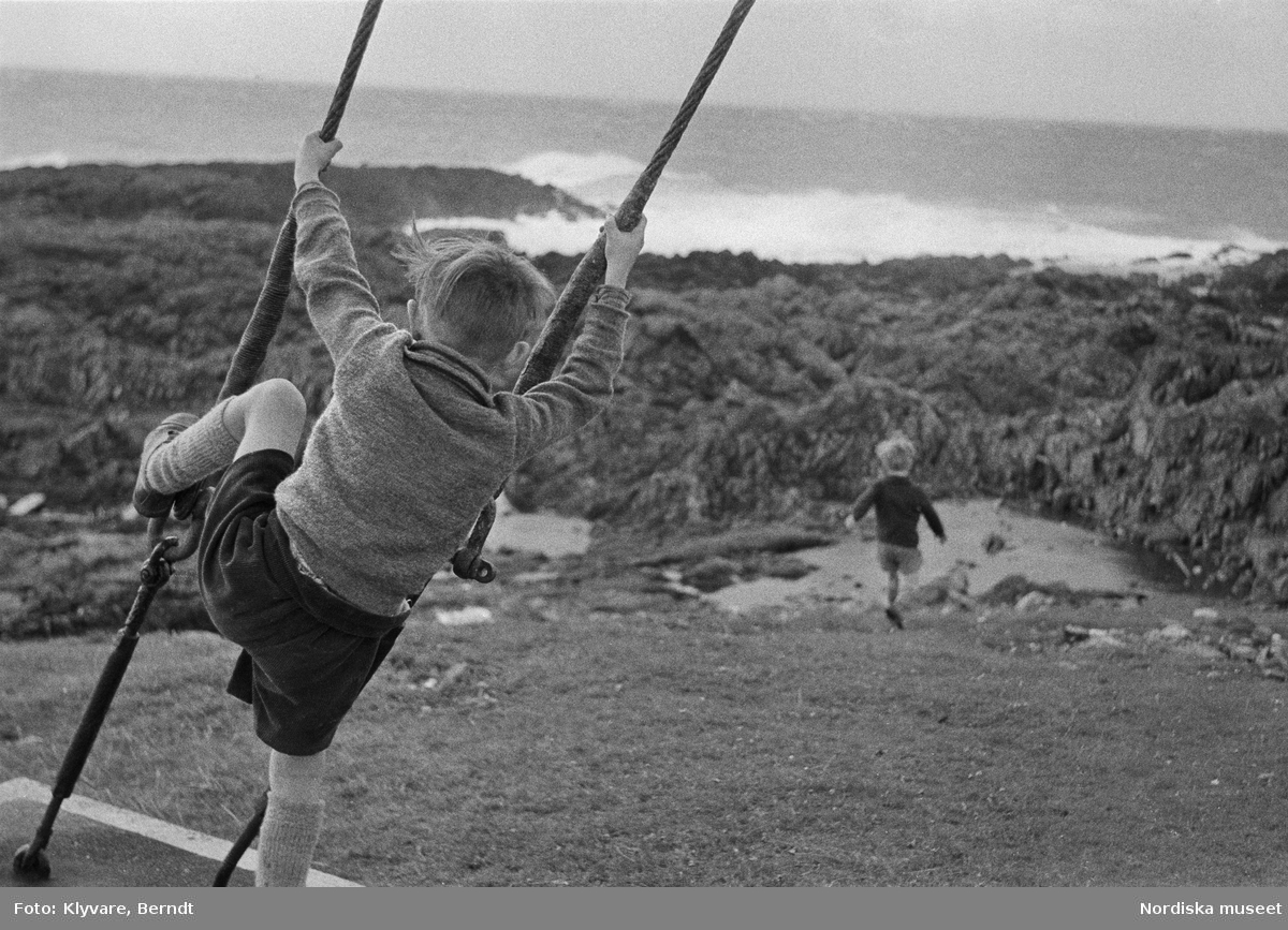 Pojke klättrar i stålvajrar en annan pojke springer iväg. Stenig havsstrand, Shetlandsöarna.