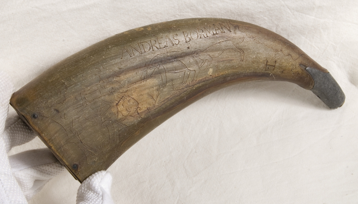 Gulgrönt horn med träbotten och tennbeslag vid mynningen. På båda sidor ristad dekor i form av jaktscener, samt namnet ANDREAS BORMAN och initialer JP. 1 [svårläst].
