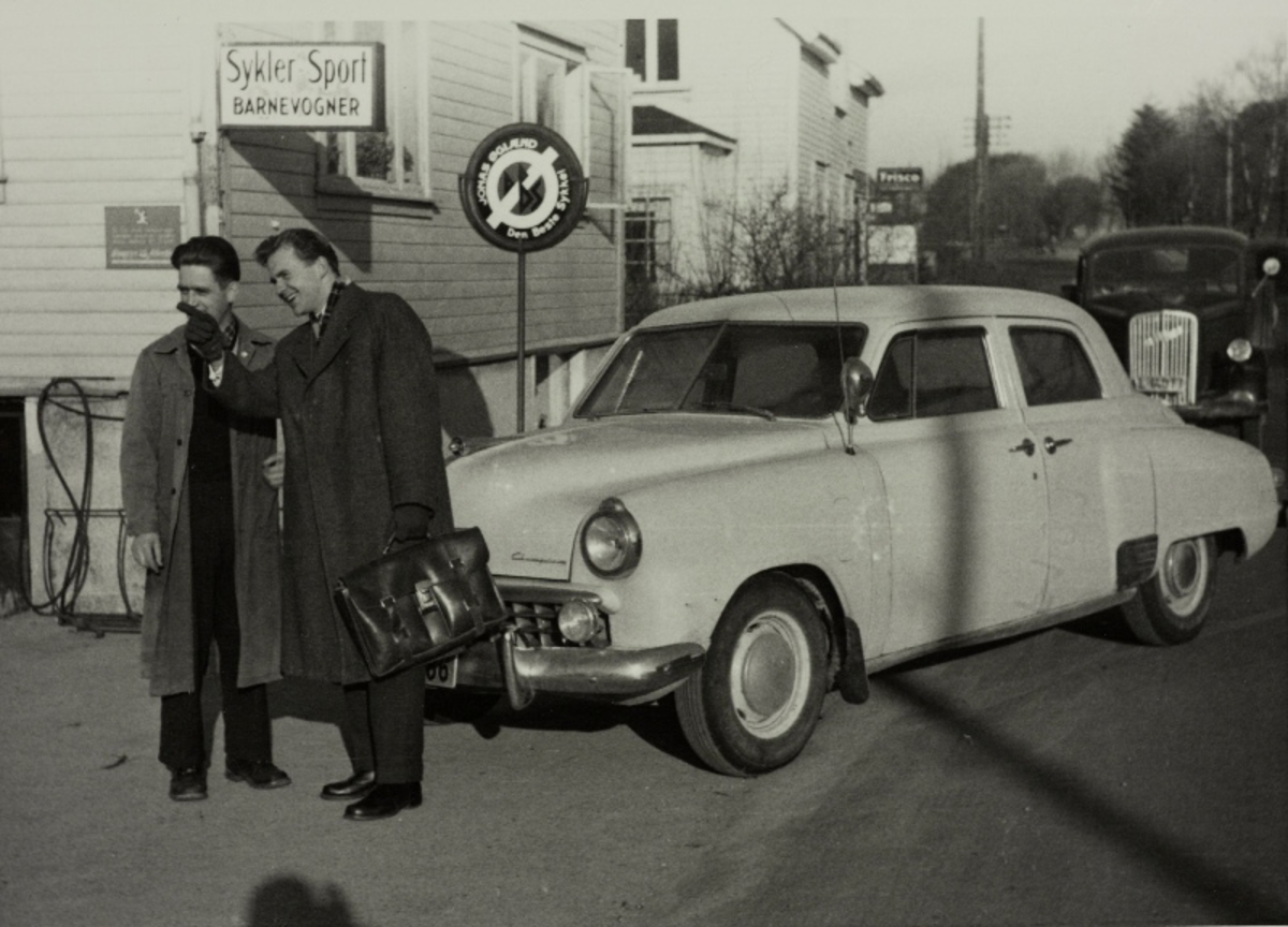 Reisende selger fra Jonas Øglænd Sandnes, sannsynligvis Øivind Øglænd, med en annen mann foran en bil (Studebaker Champion) på et gatehjørne, i bakgrunnen en Opel Blitz lastebil