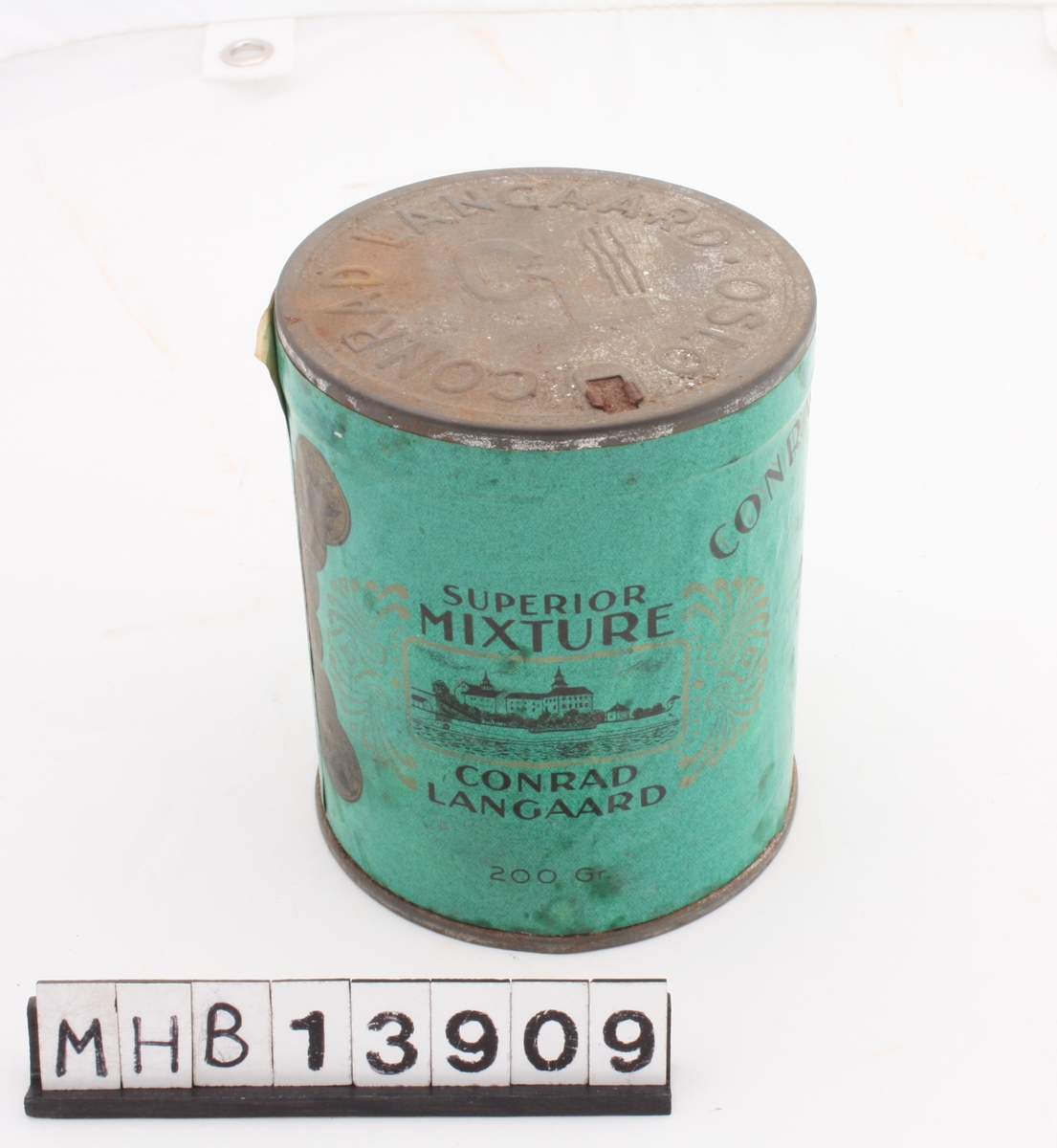 Sylinderformet tobakksboks av metall, med metallokk og papiretikette som i helhet dekker boksens stående sider.