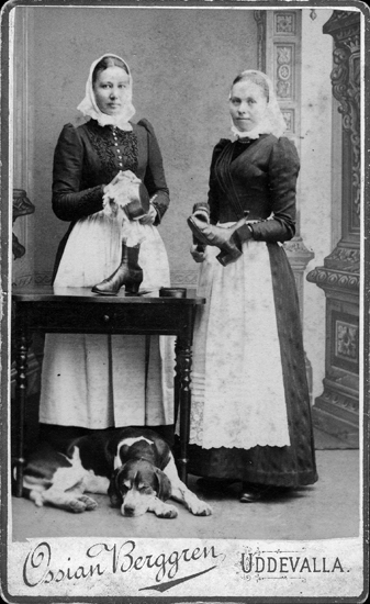 Ateljéporträtt av två kvinnor som putsar ett kokkärl respektive ett par skor, under bordet ligger en hund