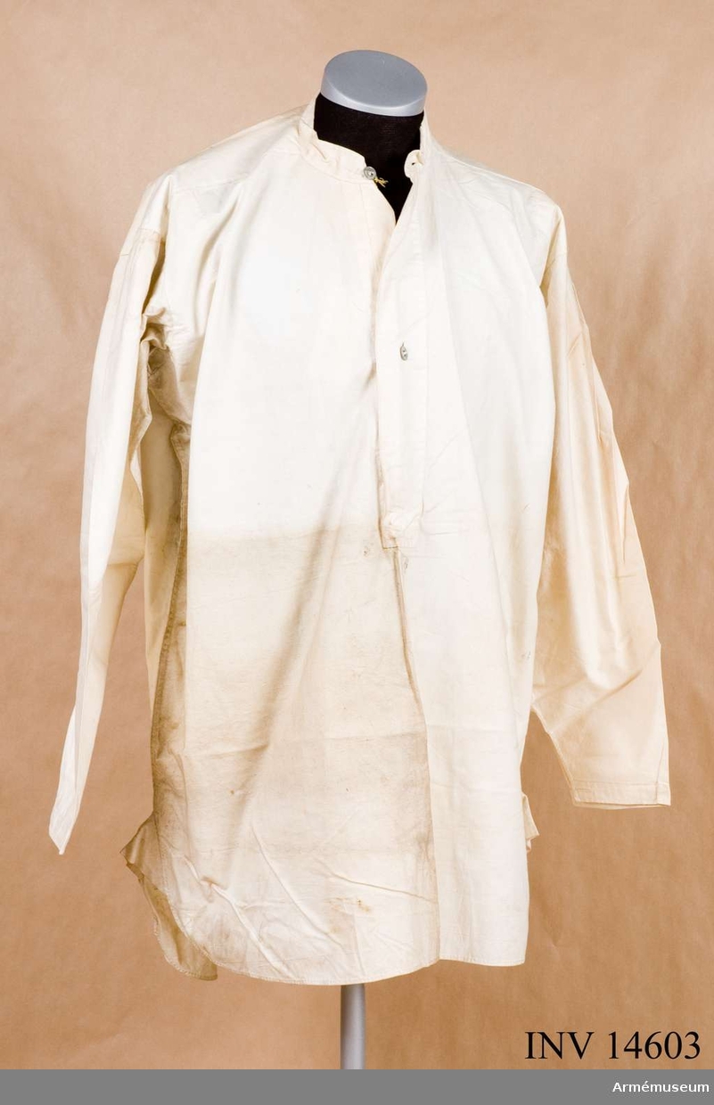 Skjorta av vitt bomullstyg med fast upprättstående krage och långa ärmar. Kragen och bröstöppningen knäppes med två metallknappar. Samma sorts knappar, en på varje ärmuppslags sprund. Skjortan lite smutsig. På skjortans framsida stämpel: "W" uppåtriktad pil "D", "6" under pilen. På kragen  påskrivning "Clara Summer 38".
