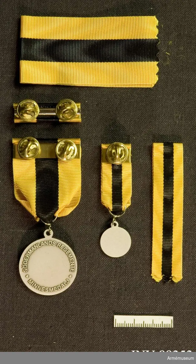 Tre samhörande föremål; en minnesmedalj, en minatyrmedalj och ett släpspänne. Band i gult med bred svart rand i mitten.