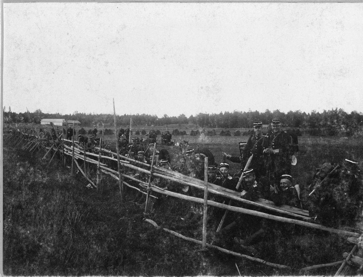 Soldater ur Livregementet till fot I 3, hukar bakom gärdesgård under manövern i Närke 1895. Soldaterna iförda uniform m/1886 och med vita band i käppin.