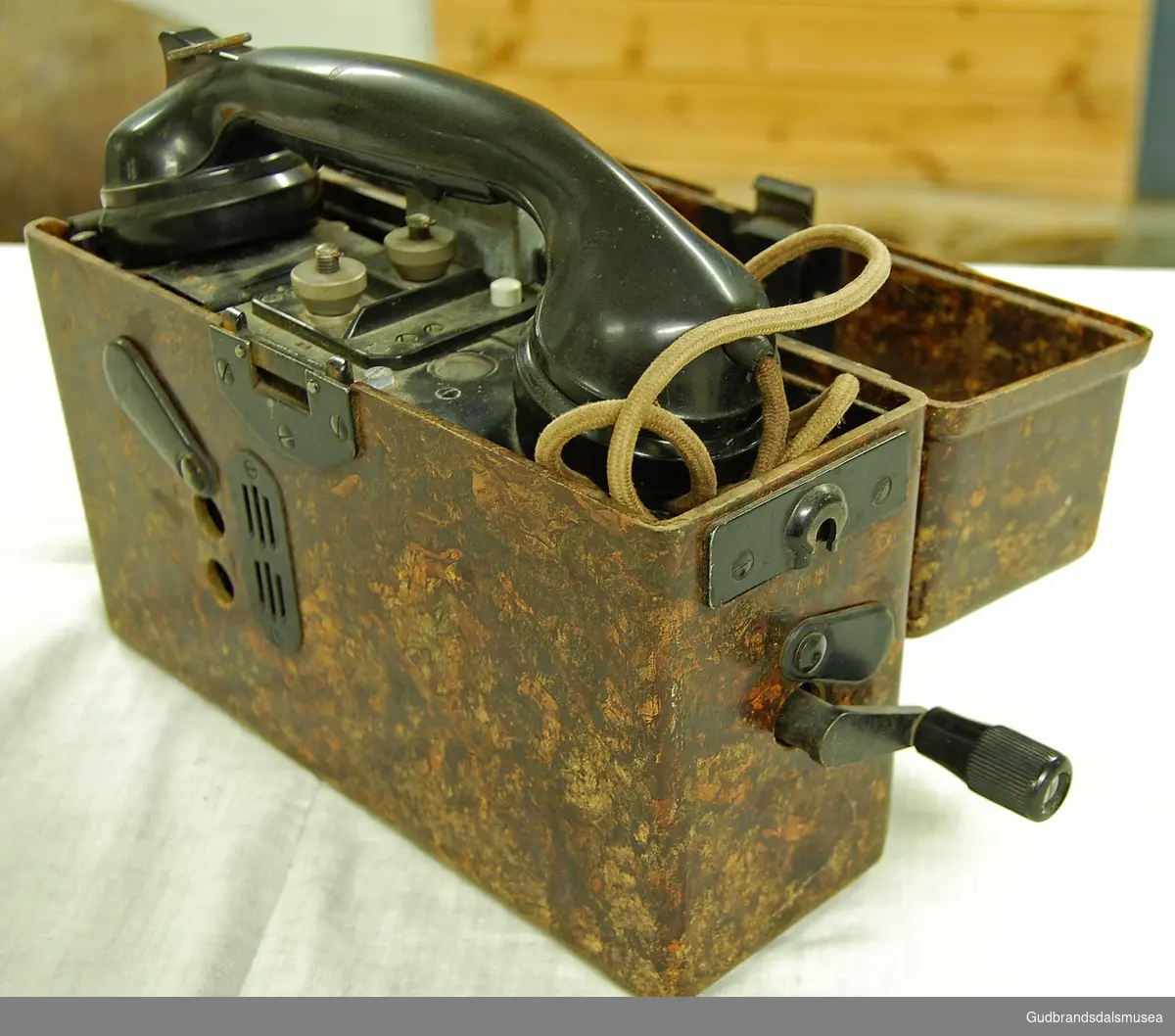 Svart telefon bygd inn i en brun, bærbar kasse. Rektangulær kasse med hengslet lokk. Med sveiv. Koblingsskjema med tysk tekst, alfabetkoder på tysk.