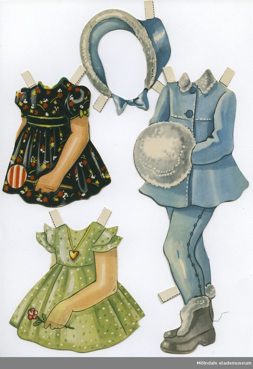 Pappdocka med kläder från 1950-talet. Docka och kläder är märkta "Karin" på baksidan - dockans namn. Dockan föreställer en flicka med blond frisyr och blå ögon, iklädd underklänning, nylonstrumpor med strumpeband och skor. Garderoben består av tre klänningar, varav en med matchande mössa, kappa och kjol med basker, samt vinterkappa med byxor, pampuscher (galosch med pälsbräm), muff och hatt. Docka och kläder förvaras i en avlång påse av smörpapper, med texten "Påklädningsdockan 'Karin'".