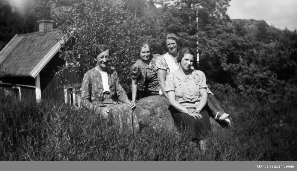 Fyra kvinnor sitter på en sten vid en äng framför en stuga. Bilden märkt: "Gäster i Kållered", 1929-1950-tal.