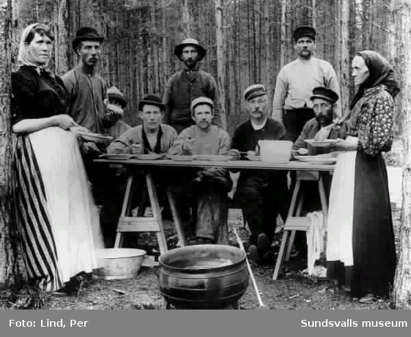 Alby 1898. Fem män sitter till bords ( två bockar och en skiva ) och äter ur djuptallrikar på bordet finns ett höganäskrus.Tre män och två kvinnor är stående. I förgrunden står ett kokkärl på ben.
