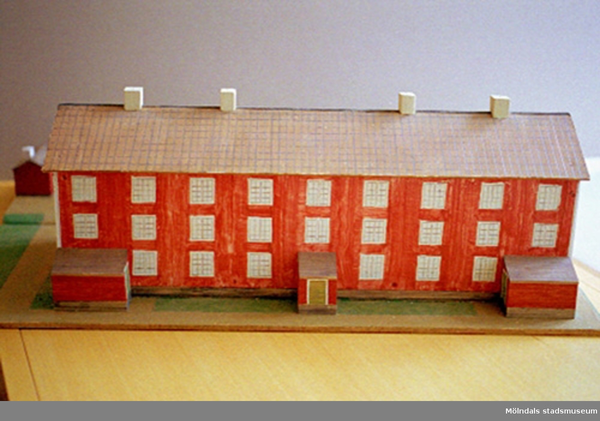 Ett rött bostadshus med tio fönster i rad, tre våningar högt, och tre entréer. På taket finns fyra skorstenar. Vid sidan av ligger två mindre stugor. Huset är en kopia av "Byggninga" som var arbetarbostäder till Anderstorps fabriker i Lindome. Se även 1994_0992 och 1994_0993. Harry Bergmans "gubbar" (träfigurer).
