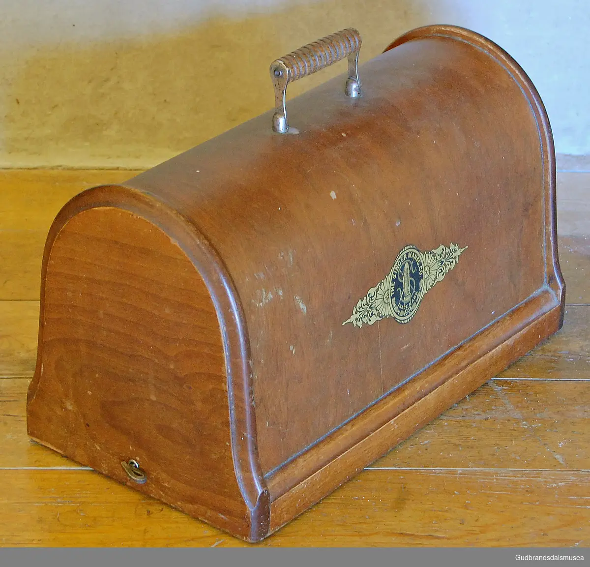 Koffert/lokk i brunt tre, til Singer symaskin, har rektangulær flate og er rund i topp. Håndtak i tre med dreide riller.  