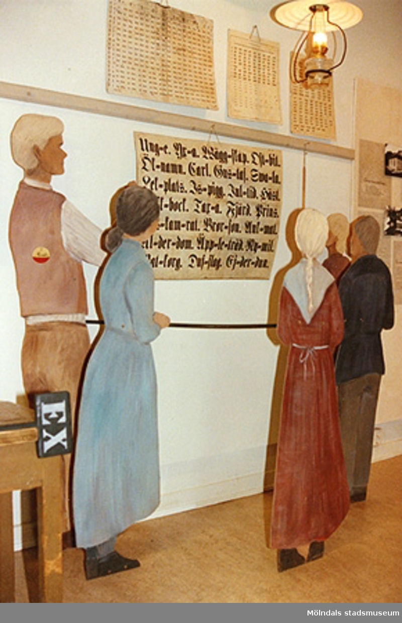 Utställningen "Lära för livet" 12 mars 1988 - 19 mars 1989 på Mölndals museum.
Fem träfigurer som tittar på en bild (med text) på väggen. Detta är en läsecirkel från en Bell-Lancasterskola.