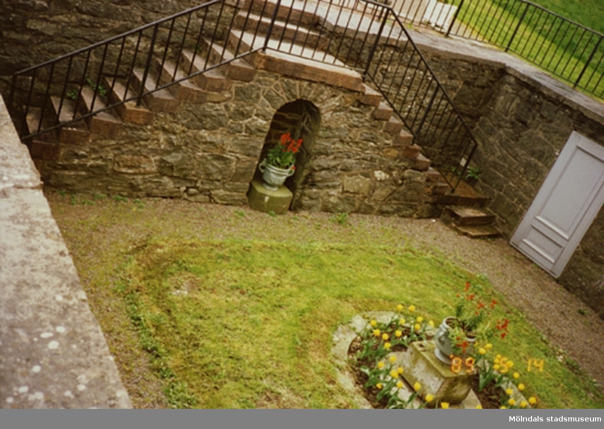 Den nedsänkta innergården (Köksgården) med gräsmatta och blomurna i höger nederkant. Innergården står på Gunnebo slotts östra kortsida (östra parterren). I bakgrunden står en krukväxt placerad i nischen, som är placerad mellan två trappor.