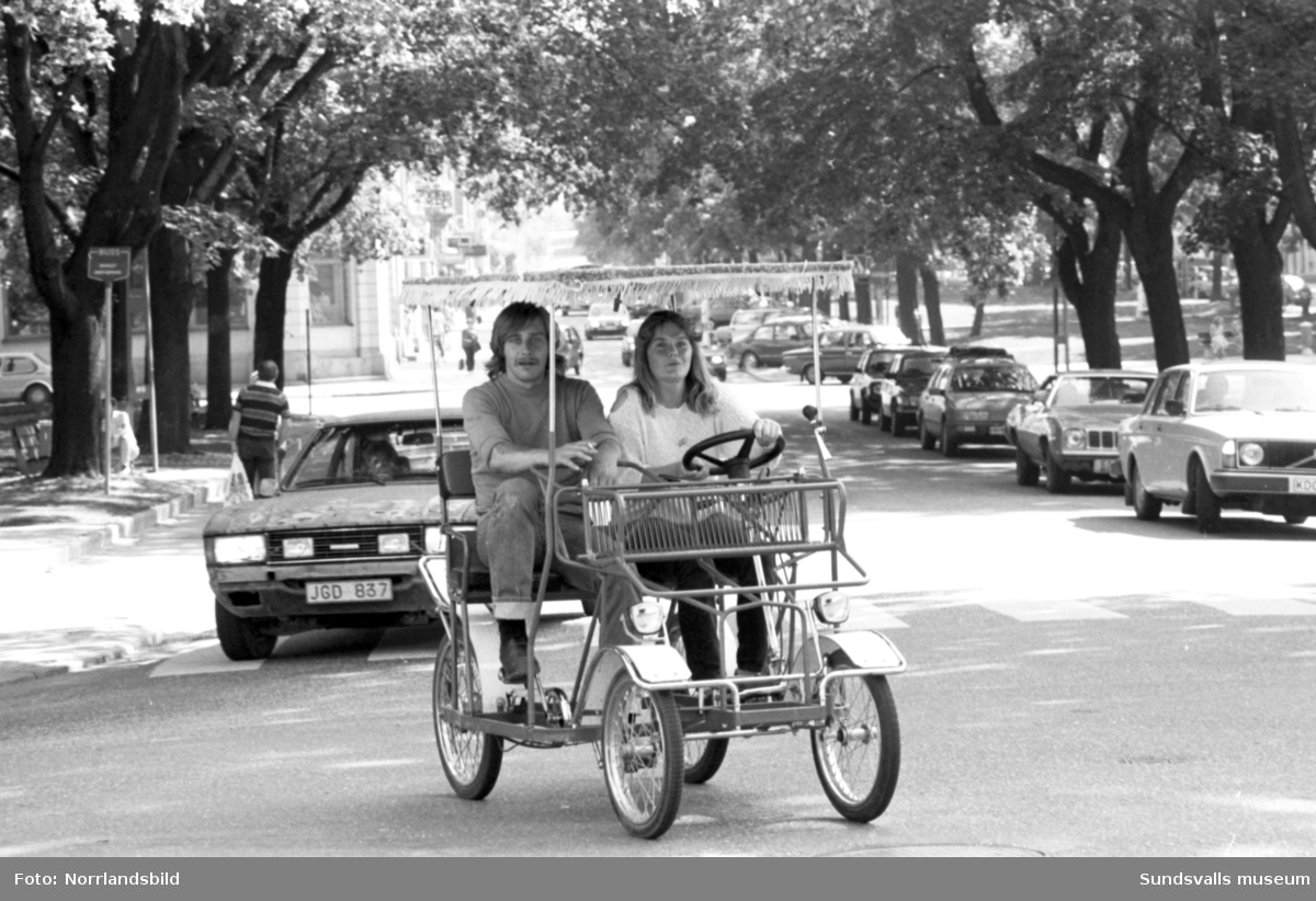 "Cykelbil." Ett ungt par framför en slags fyrhjulig cykel med tak och ratt på Esplanaden.