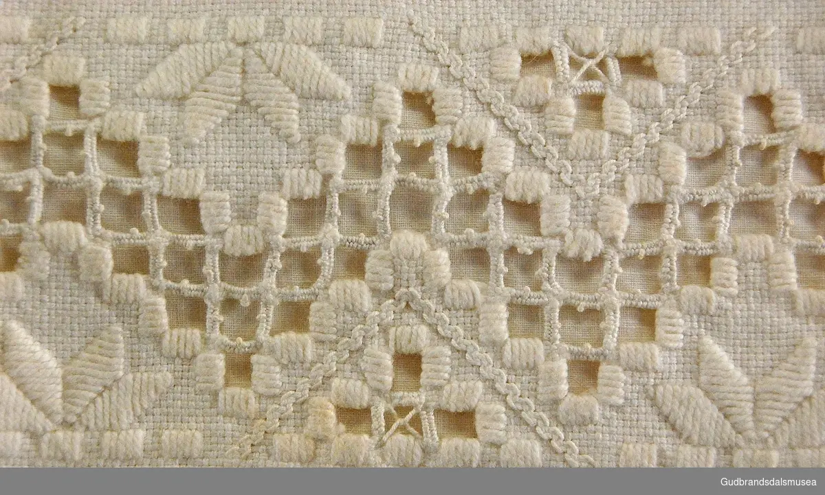 Hvitt, rektangulært putevar med brodert/sydd  innslag; hullsøm, sikksakk-mønster, halve kronblader. Med lukking på ene langsiden; to knyttebånd på hver side.