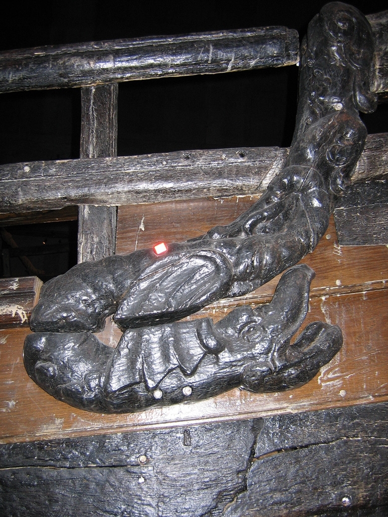 Skulptur av en drake återgiven i höger profil.
Draken har ett stort huvud med kraftigt uppdragen nos och öppen käft. Drakstjärten och vingen övergår i den ovanliggande tritonen, se fyndnummer 23059. Baksidan är slät.
Skulpturen är välbevarad.

Text in English: A sculpture of a dragon in right profile.
Large head with very upturned nose and open jaws. The dragon''s tail and wings change into a Triton that is situated above, see No. 23059. The back is smooth.
The sculpture is well preserved.