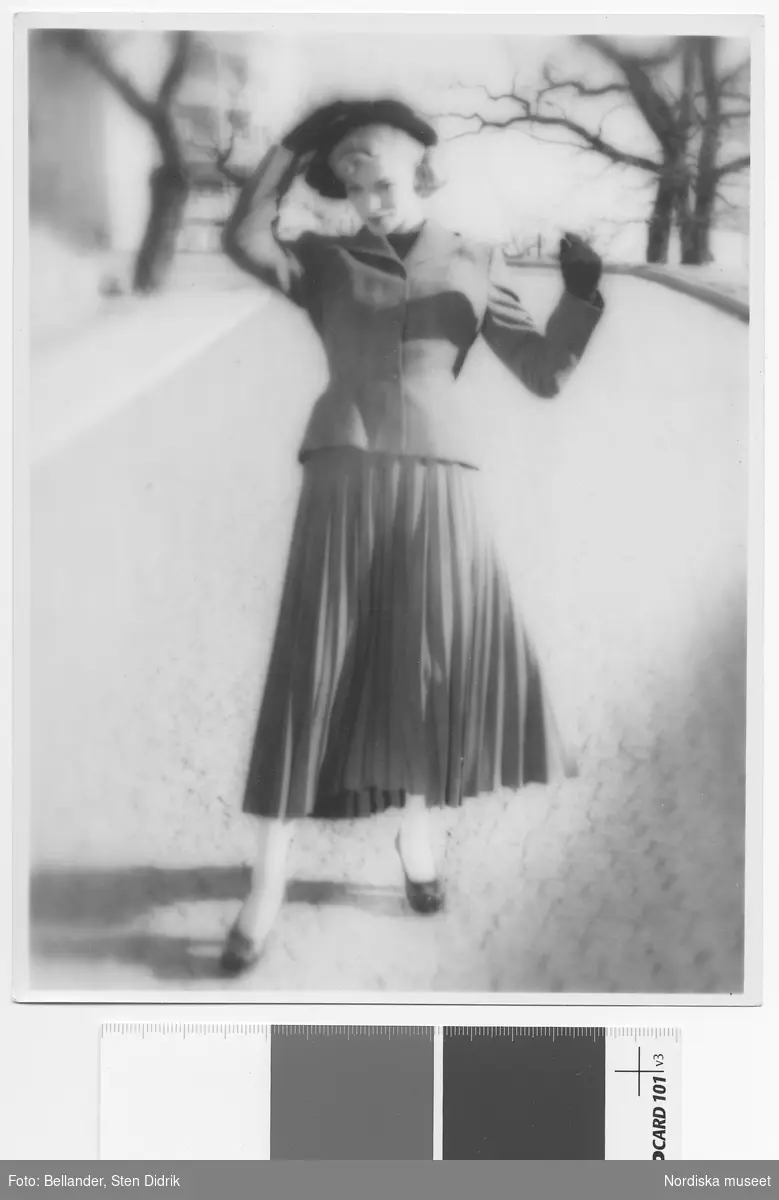 Modell i jacka och plisserad kjol, hatt och handskar, går på kullersten.