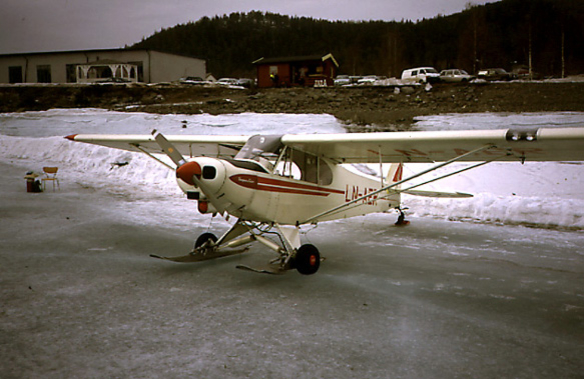 Lufthavn, 1 fly på bakken, skrått forfra. LN-AER "Super Cub", Piper Aircraft Corporation PA-18-150, fra Valdres Flyklubb. Med ski montert på hjulunderstell. Bygning bak. Snø på bakken.