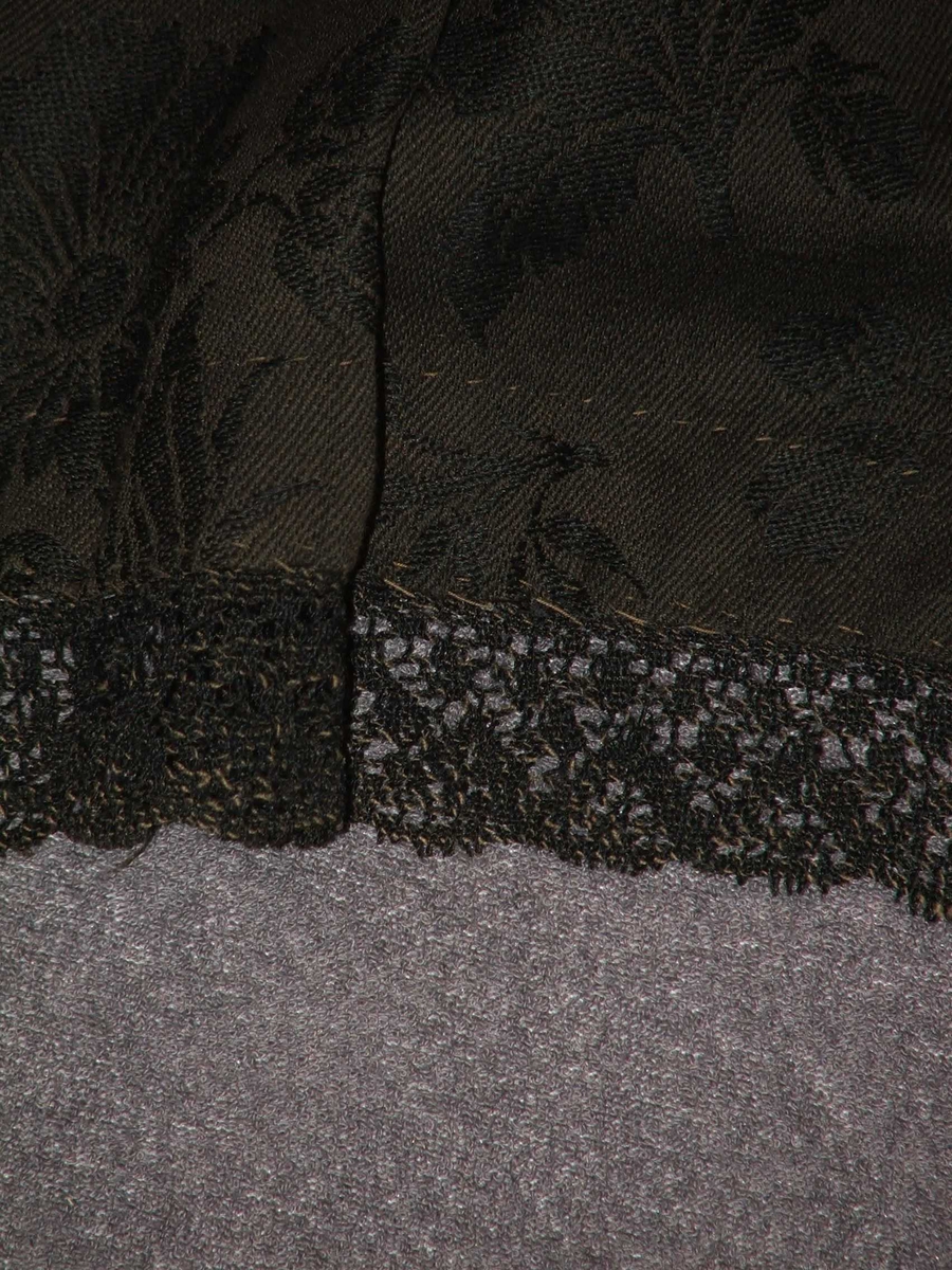 Forkle, svart med innvove blomemønster i møskbrunt. 3 cm. maskinblonde nede. 2 cm  skodning (kanting på vranga nedst) i raudmønstra bomullstoff.Plissèfalder i livet og  hekter på kvar side. 
