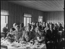 Gruppe menn og kvinner i sal med festpyntet bord. ?? inne i 