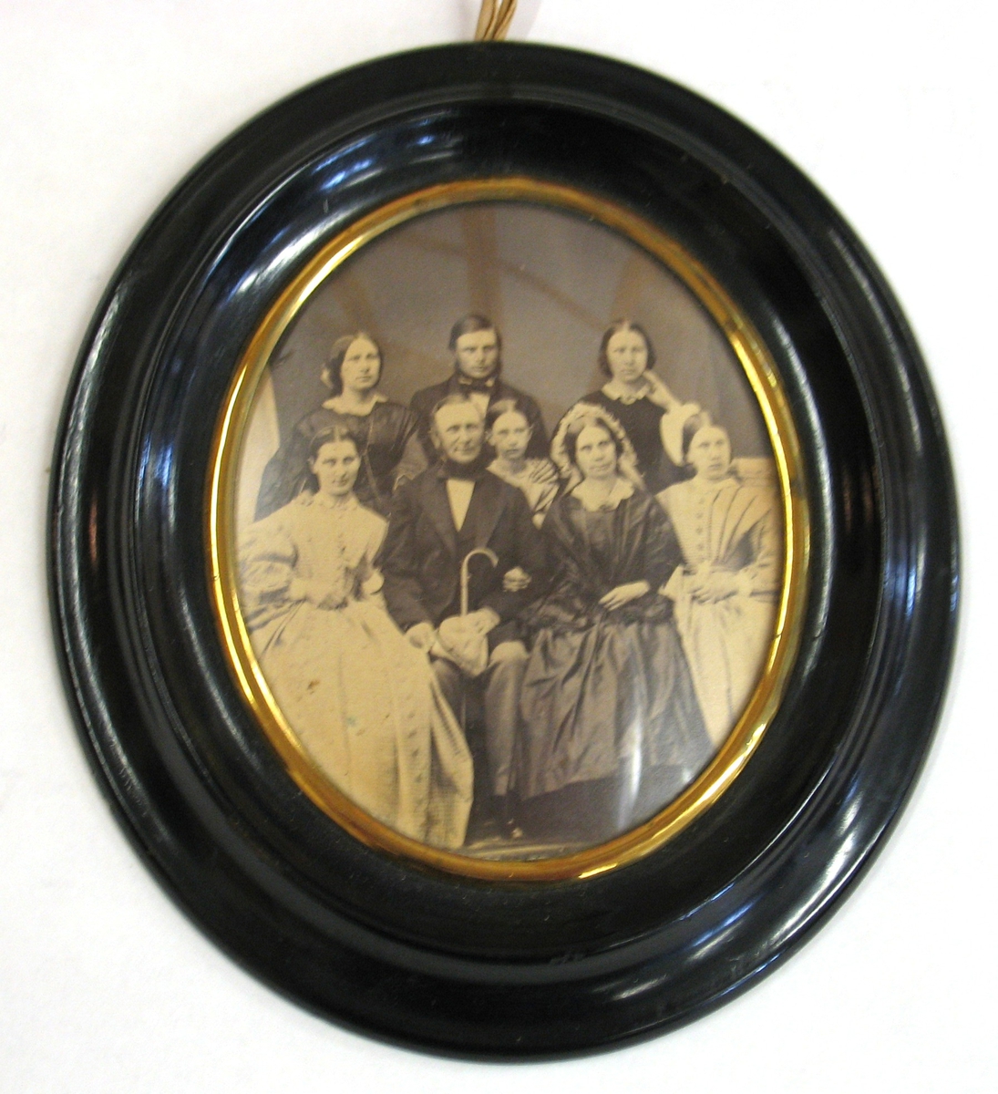 Motiv/dekor: Familjeporträtt (8 personer).
Svart oval ram med guldlist.
