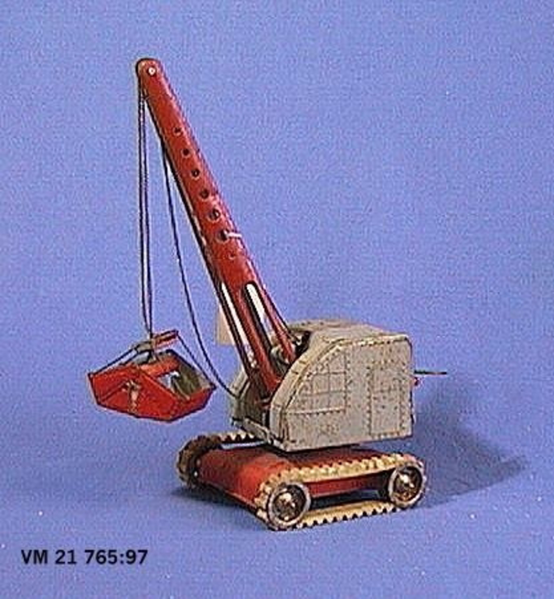 En grävskopa på larvfötter tillverkad av plåt. Skopan är löst hängande i en kedja som kan styras från förarhyttens baksida. Tillverkad av MFZ i U.S.-Zone, Väst-Tyskland, under slutet av 1940-talet.

L. 30,5 cm.  B. 13 cm.  H. 11,5 cm.