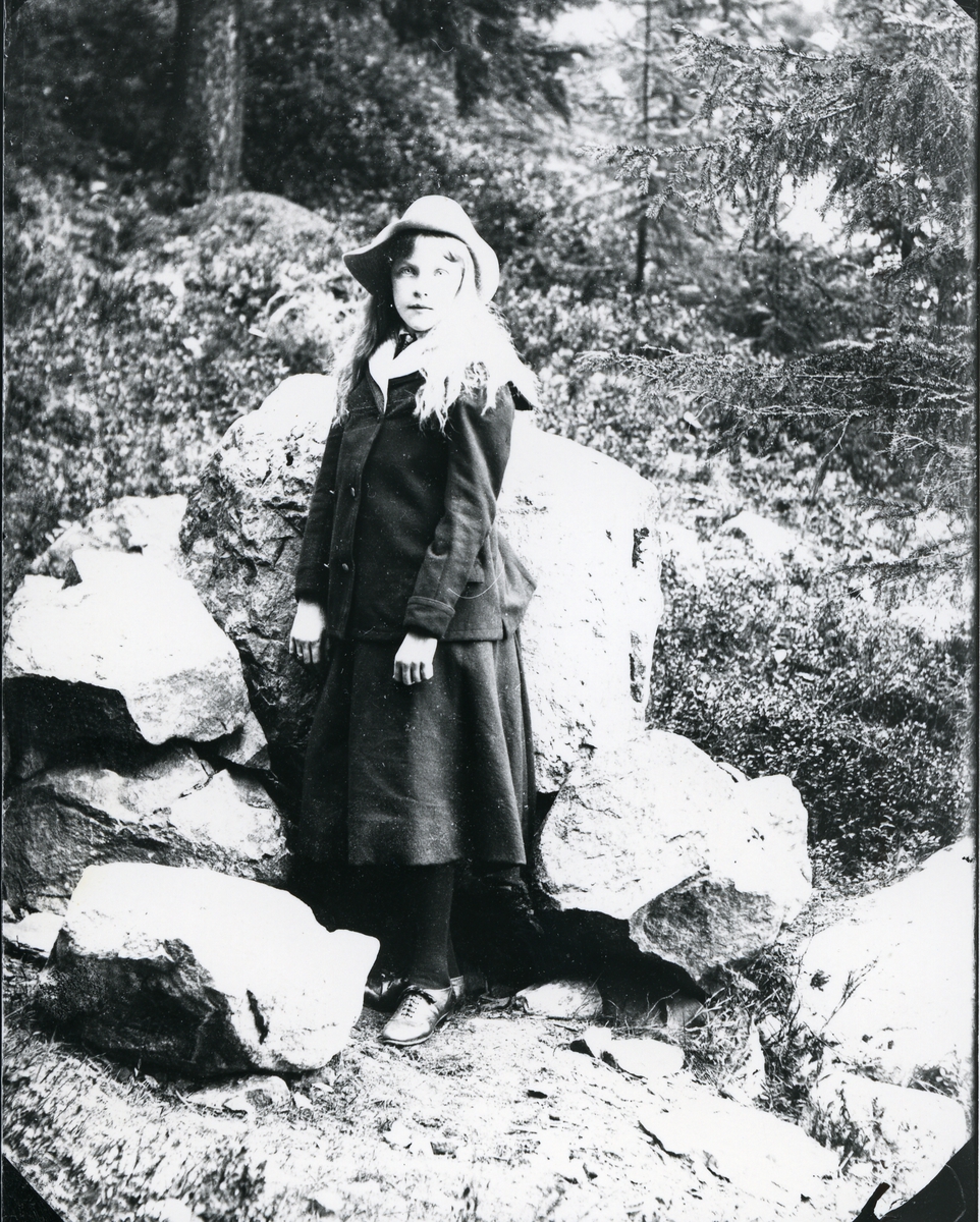 Ung jente med hatt, i helfigur, foran store steiner i skog