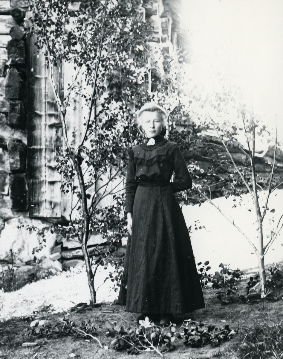 Ung kvinne i helfigur, kledd i mørk kjole med sølje, avbildet foran låve og låvebru