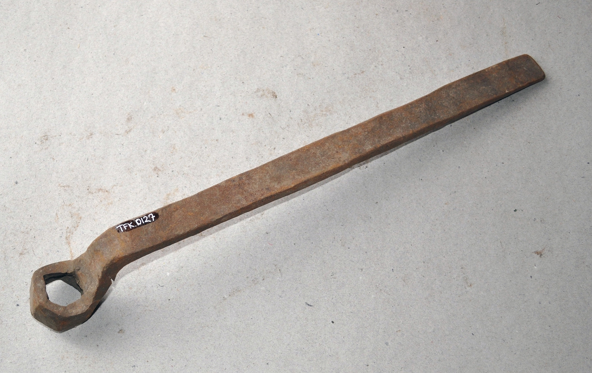 Håndsmidd sekskant fastnøkkel av jern, med 1 1/2" lukket kjeft. Langt håndtak av jern, med en bøy på halsen. Fastnøkkelen er smidd av et flattjern.