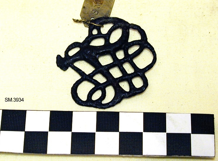 Fra protokollen: Bronsebeslag i urnes-stil, som Shetelig: Urnesgruppen, fig.24, i årb.1909. Stykket består av en en dyrefigur i oppløst, gjennombrutt slangeornament, og er så og si identisk med typeeksemplaret, bare få og uvesentlige avvikelser, idet deyrets hode og hale er litt annerledes utformet, videre har vårt typeeksemplar avbrutte små tagger på undersiden der hvor typeeksemplaret har huller. Til og med overflaten er ornert ens, med små tverrstreker på dyreslyngningene. Stykket er meget godt bevart, fargen er mørk, nesten svart.

Funnet på kirkegården, ellers ingen opplysninger, men må vel forstås som reminisensen av hedensk gravskikk i tidlig kristen tid.
Datering: Slutten av 11. årh., mulig begynnelsen av 12 årh.