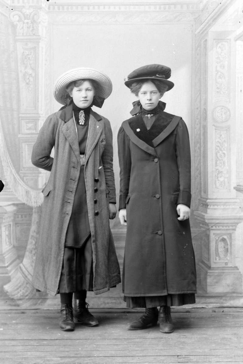Studioportrett av to kvinner med frakk i helfigur.