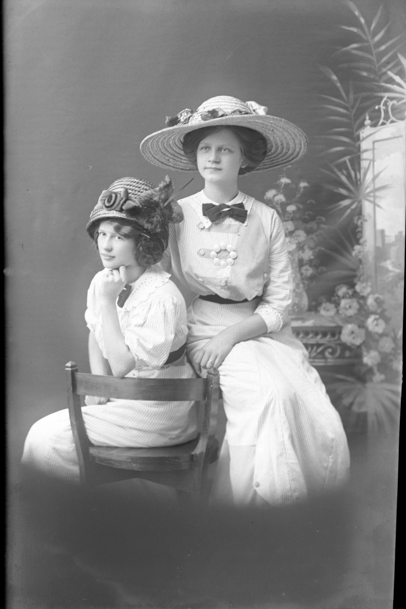 Studioportrett av to kvinner med blomster på hattene sine.