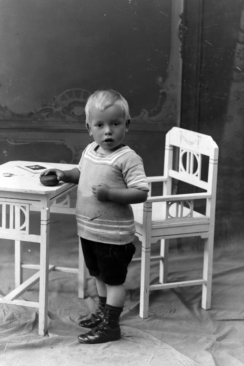 Studioportrett av en liten gutt ved et bord og en stol.