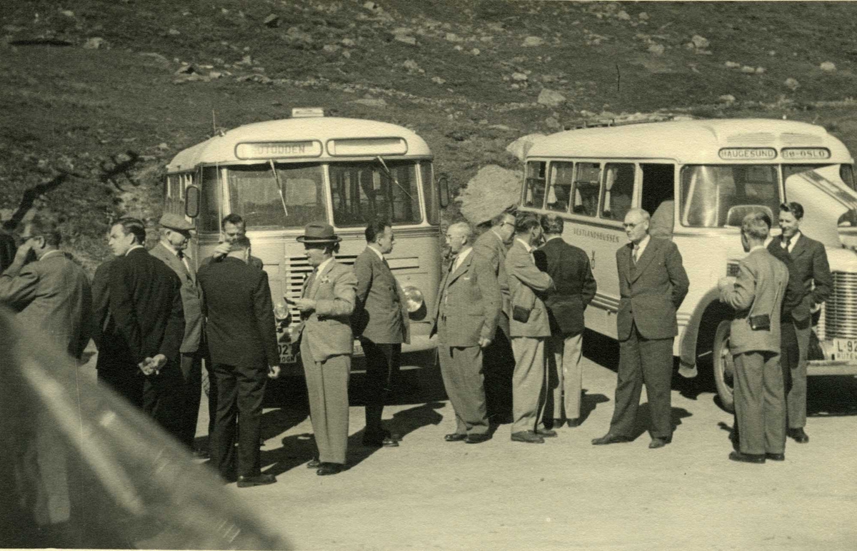Haukelisæter 1952. Jernbanekommisjonens befaring. Fjorten menn står og samtaler foran to parkerte busser. Steinet landskap i bakgrunnen.