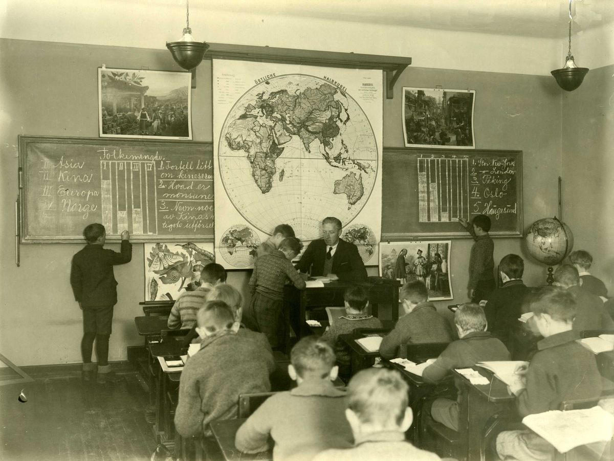 Klasserom - geografitime. To elever står oppe ved tavla på hver side av kateteret. To elever står ved kateteret der læreren sitter. Lærer er Styrskår Vik.