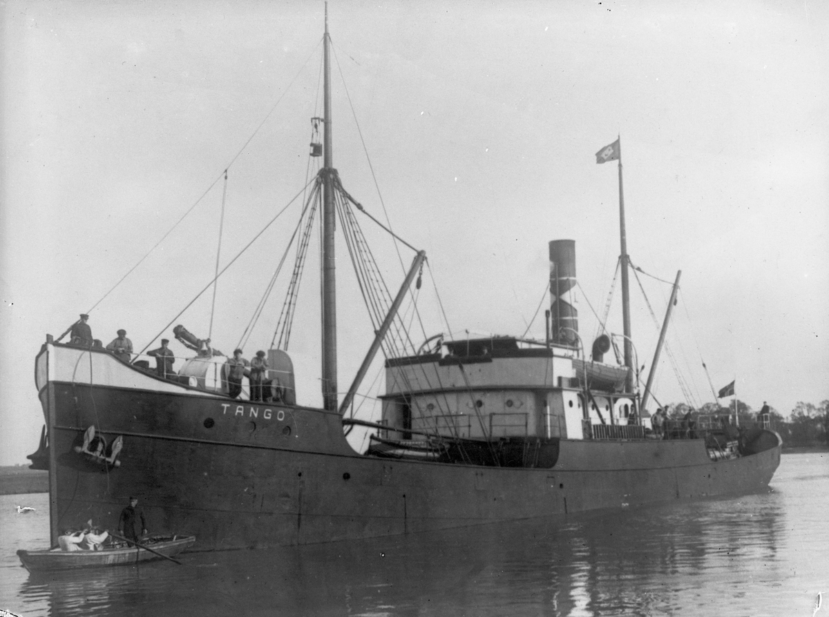 Dampskipet D/S "Tango" ved land. En robåt, rodd av to personer, ligger ved baugen og det står flere menn på dekk.