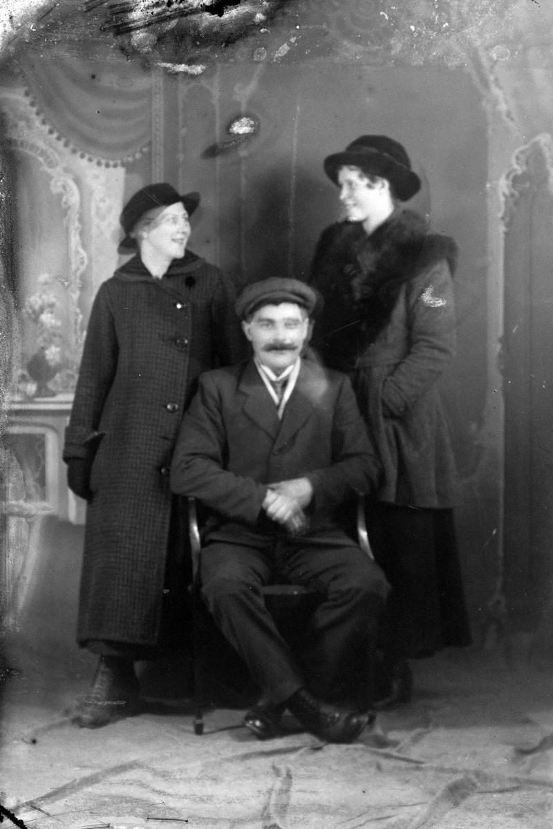Studioportrett av en mann sittende foran to kvinner som står.