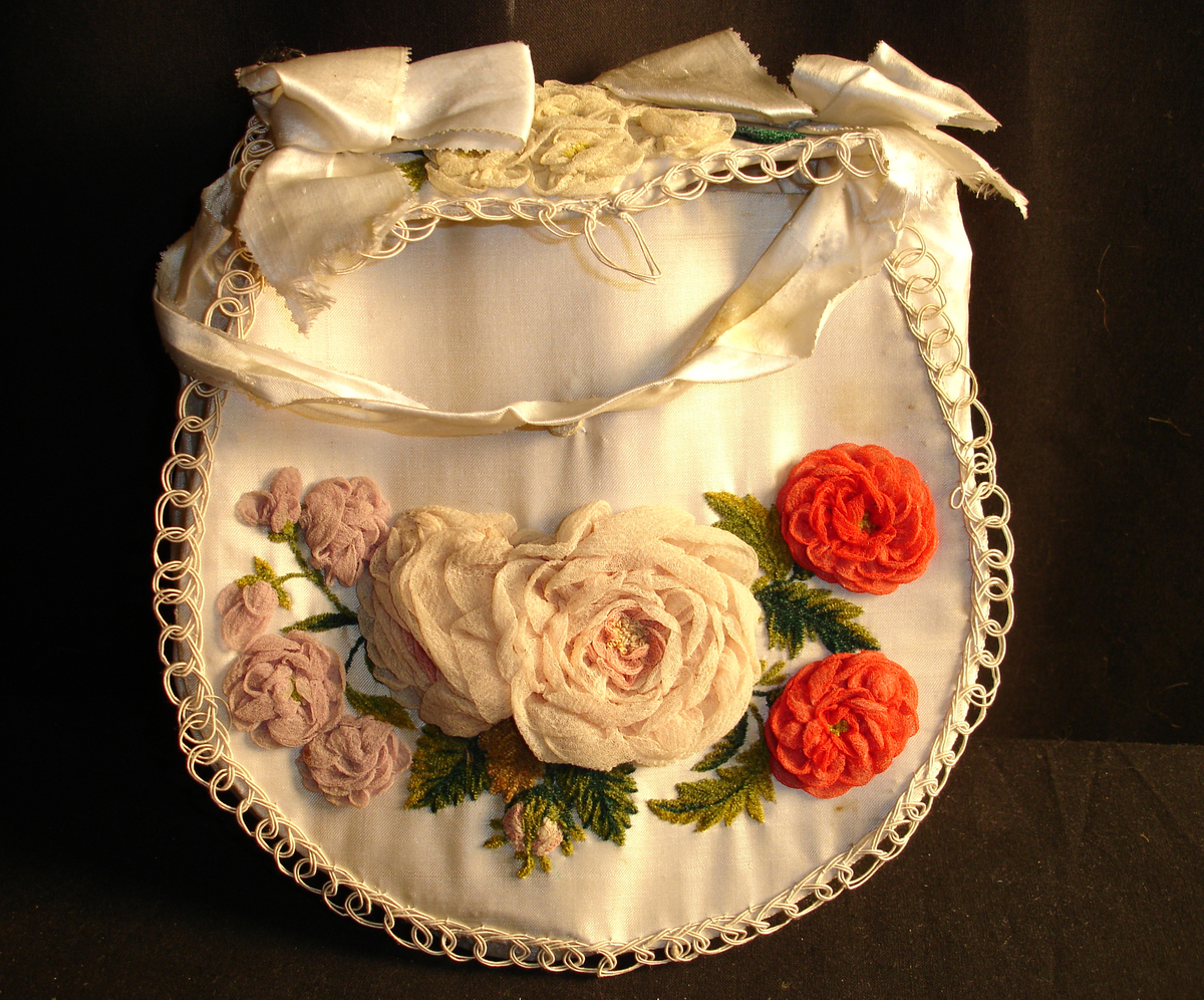 Veske i silke dekorert med blomst og broderier. (er slitt på sløyfen)
Form: oval
Tilhører brudekjolen KMK.05523
