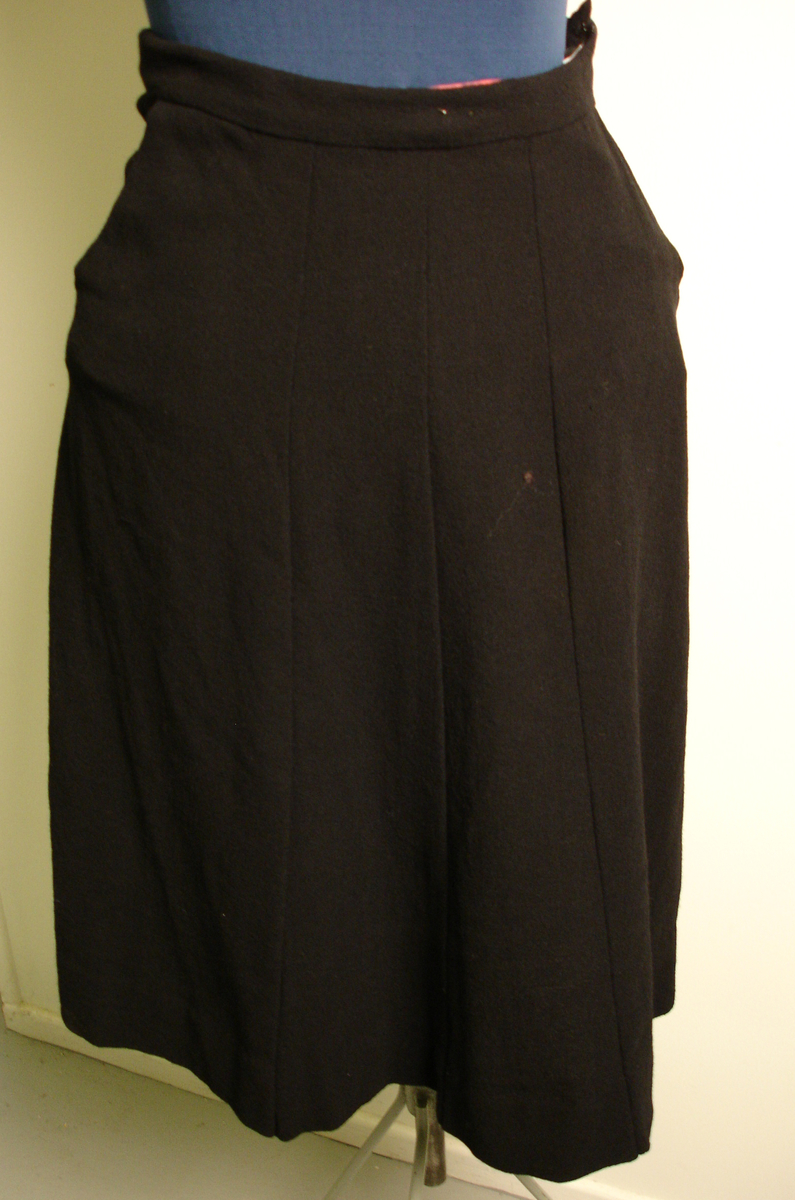 Skjørt, svart i ull, med glidelås, antagelig fra etter krigen, knekort, sømmer sydd slik at de danner en form for folder. 