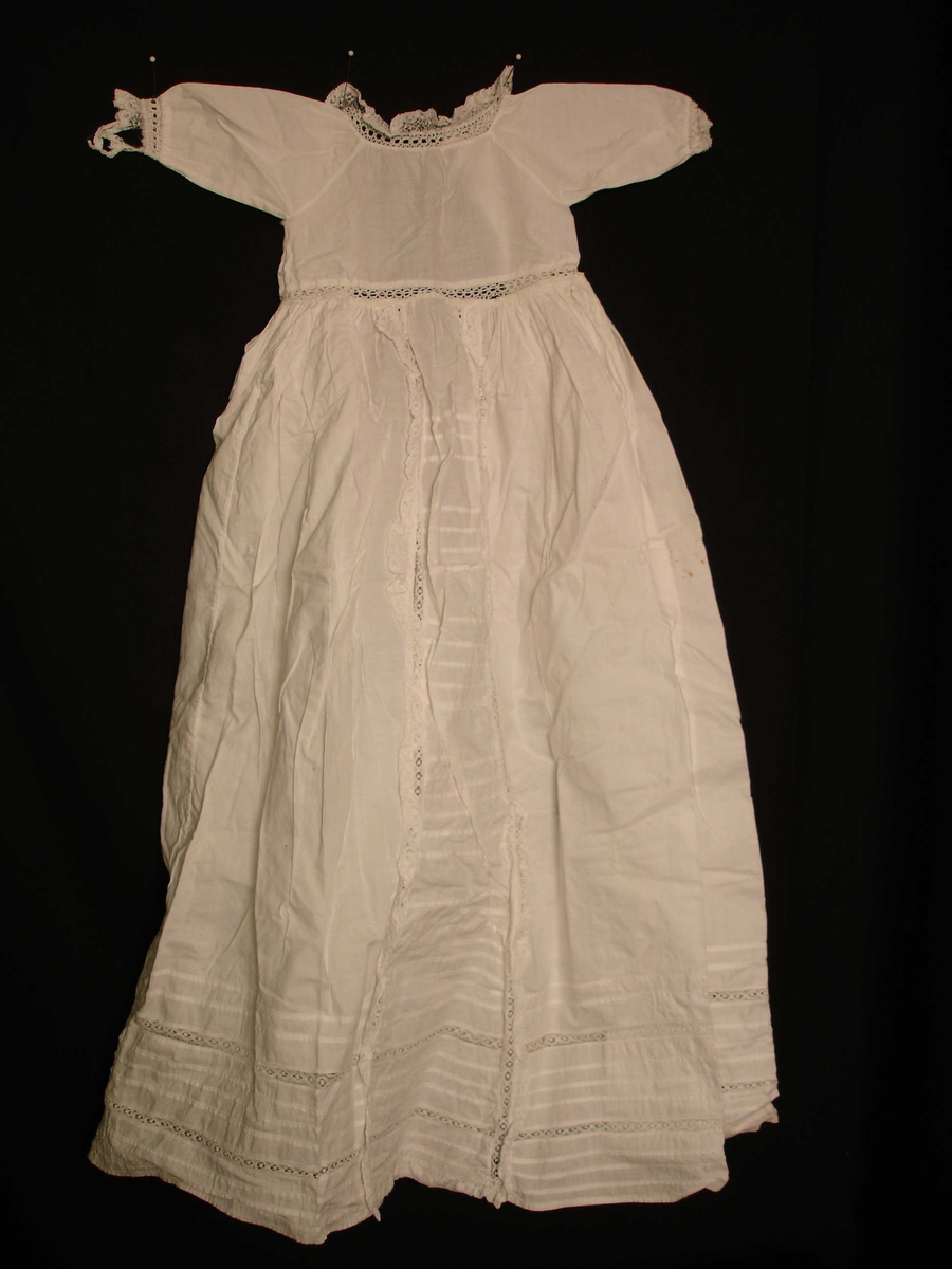 Dukkeklær, kjole i hvit bomullstoff,  blonderkant rundt hals, langs front fra midjen og ned, og ermeåpning. 