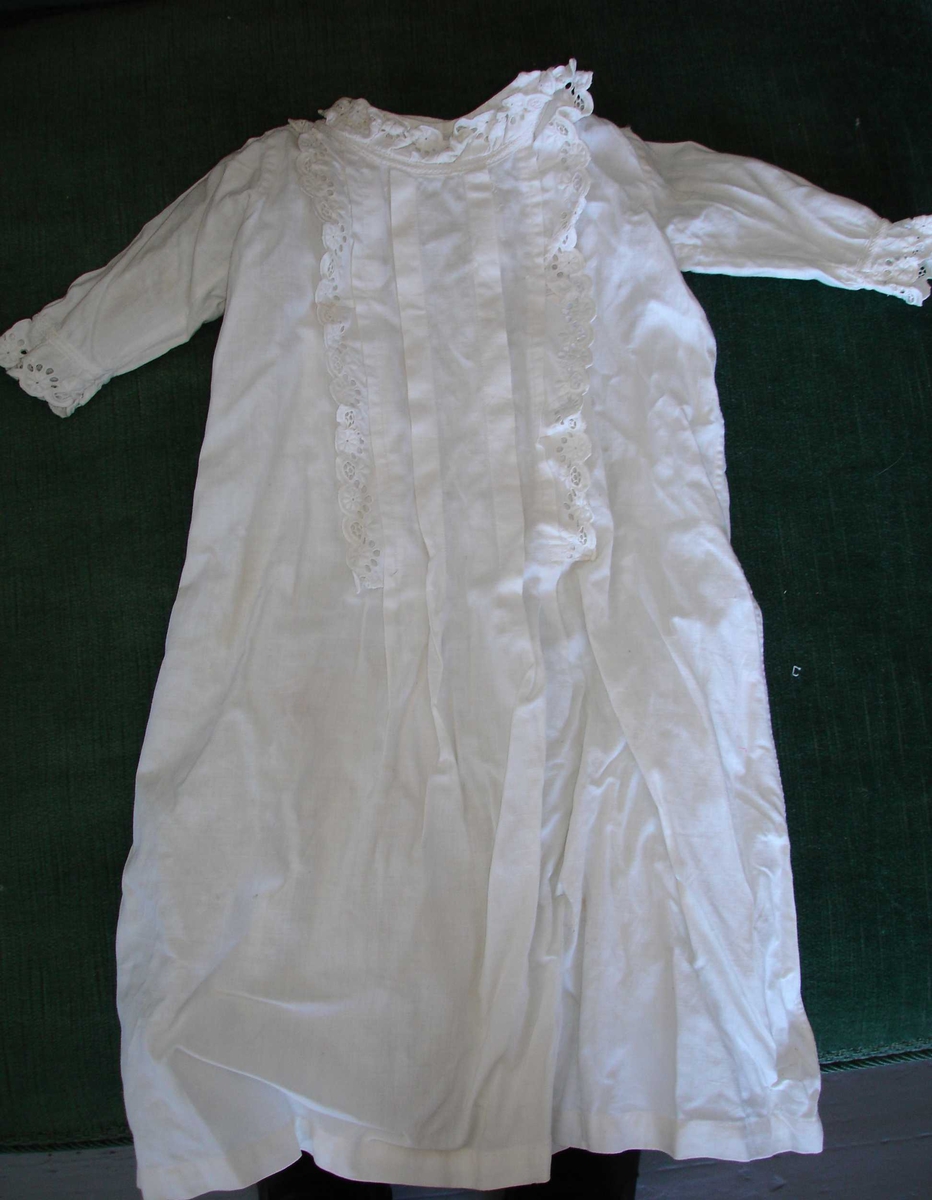 Dukkeklær. Kjole i hvit bomull med blondekant som kappe rundt halsen og langs kanten foran, samt nederst på ermene. 