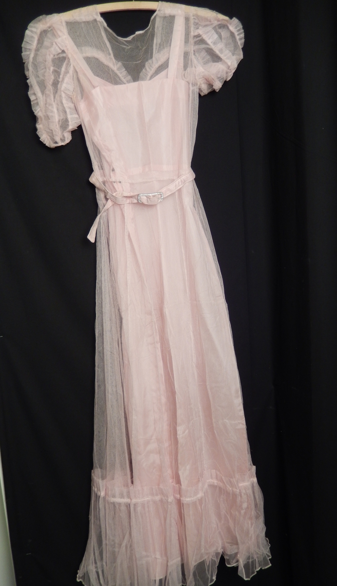 Kjolen ser ut som hjemmesydd. Men er en et merke 38 bak på kjolen, som er størrelsen. Kjolen består av to deler, underdelen av rosa nylonstoff, stropper ved skuldrene. Ytterdelen er i rosa chiffong med kappe påsydd på nederte delen av kjolen. Smalt belte i stoff med sølvfarget spenne. Rysjer på ermer og på bryststykket. 
Hatt formet som en slags bøyle, med rosa chiffong og nylonstoff festet på stålbøyle med strikk til feste bak. hatten eller hårpynten er med blått nettingslør. merkelapp Erna Marguerithe Oslo