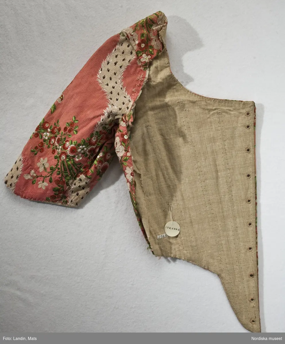 Del av klänningsliv, kvinnlig dräkt, Ängelholm (Skåne), 1760-80. Siden.
Söndersprättade delar till en ofullständig klänning. Tyget en sidenbrokad på stark rödrosa ripsbotten med blombuketter i vitt, mörkrött, rosa, och två gröna nyanser och slingrande vågband i vitt med mörklila prickar. Höger och vänster framstycke med isydda korta ärmar. Flera skarvade bitar. 10 par snörhål i framkant, foder av oblekt linnelärft, i ärmarna vitt linne. 2 avlånga bitar som varit hopsydda på mitten. I var ände sitter delar till livets bakstycken. Märken efter breda lagda veck längs ena kanten. På båda delarna fuktränder och blekning där de varit hopsydda. Svängda stycken med en rynkad sida. 4 bitar av ärmarnas engageanter.
Tyget som är från 1760–70-talet verkar ha återbrukats flera gånger, möjligen inte bara som klänning utan även som möbelklädsel (de smutsiga och blekta delarna kan tyda på det).