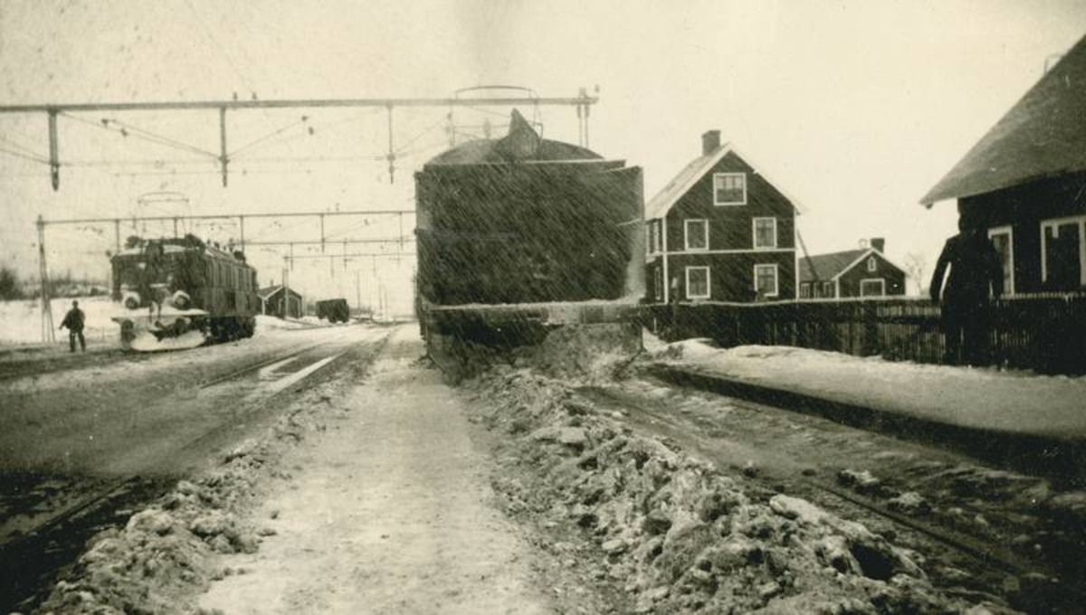 Jernbanestasjon med et lokomotiv til venstre. Bygninger.
Roterende snøplog som fjerner en snø-rygg som lokomotivet med plog har samlet midt i sporet.